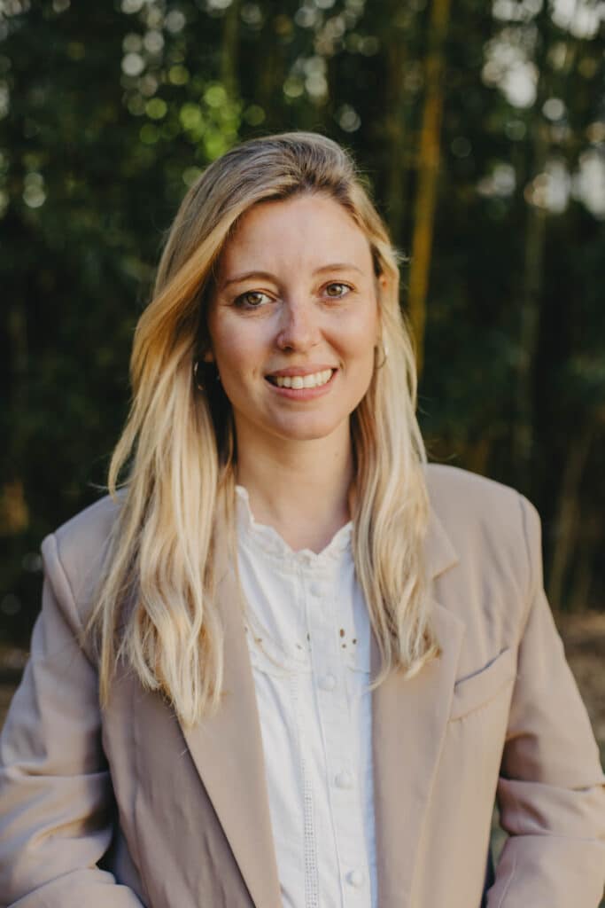 Portrait d'une femme blonde souriante en blazer beige Femme blonde souriante en blazer beige et chemisier blanc devant une forêt floue en extérieur.