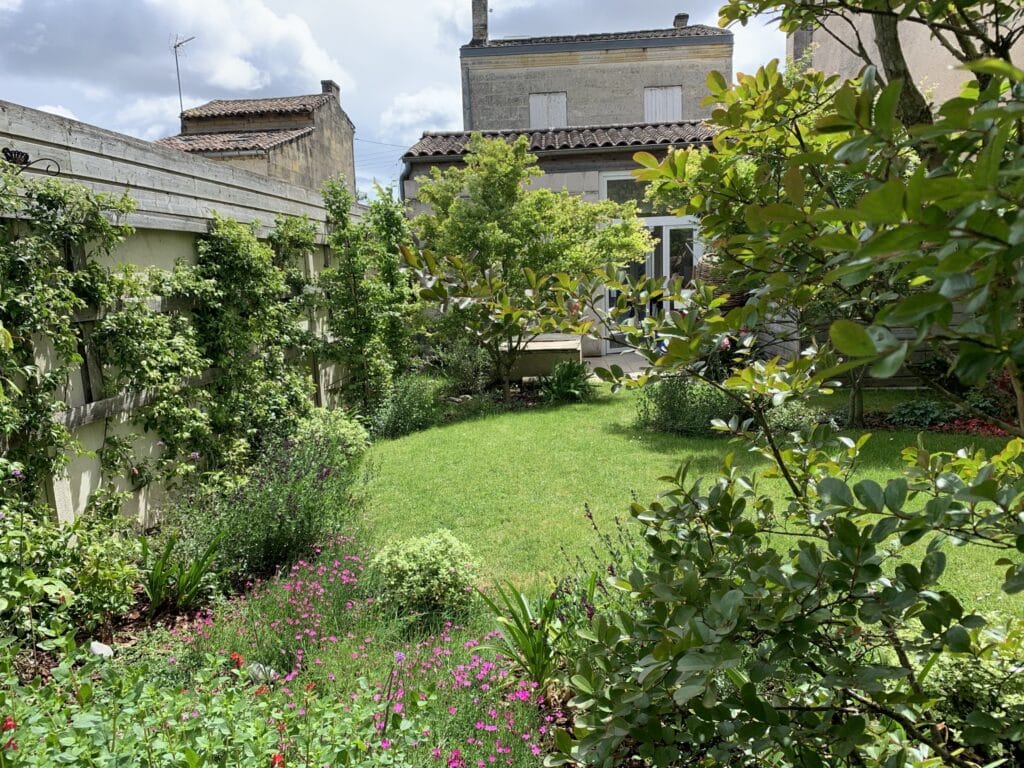 Jardin verdoyant avec pelouse, fleurs roses et plantes grimpantes bordé de maisons anciennes en pierre.