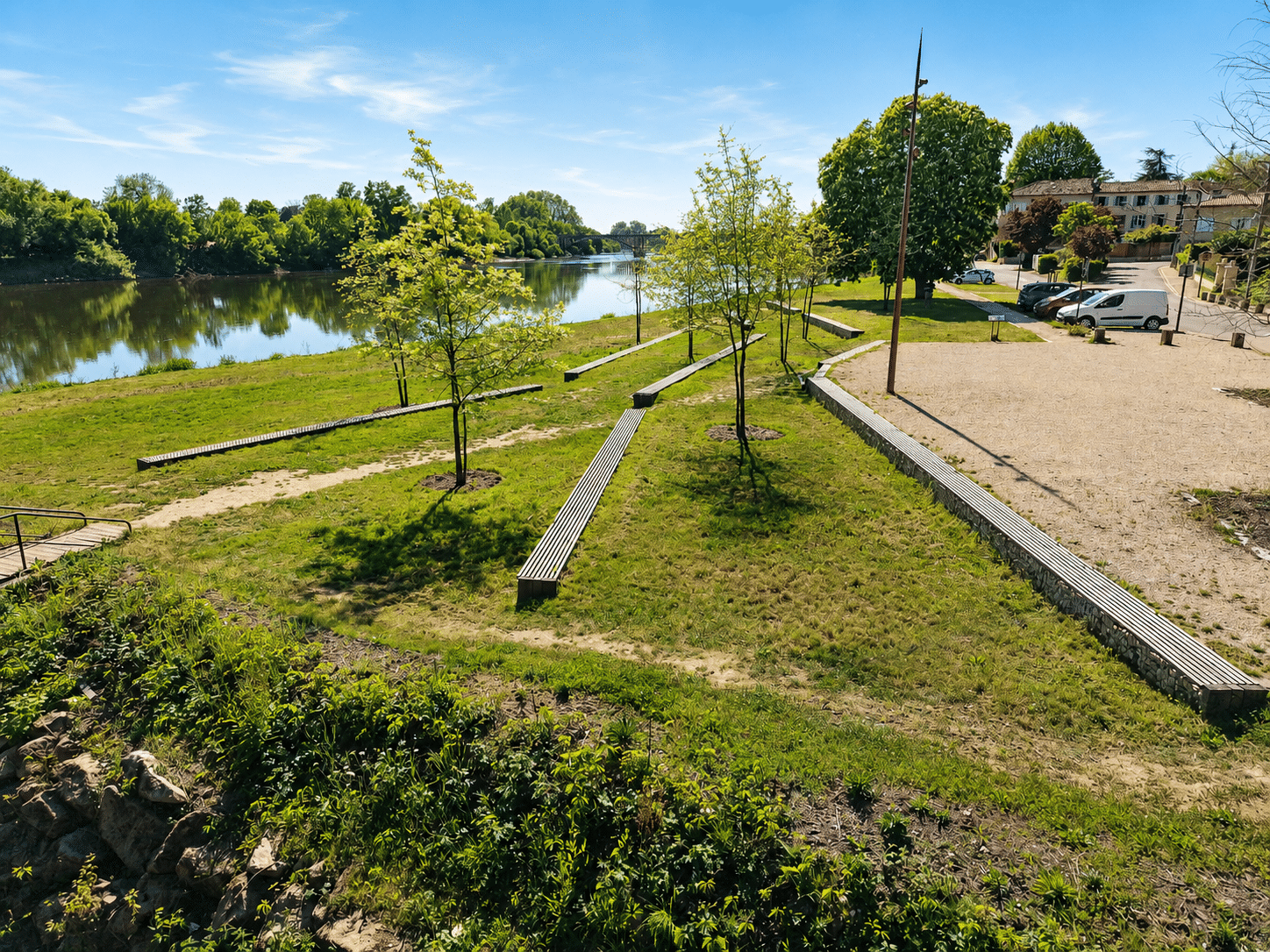 Parc paysager au bord de la rivière avec bancs en bois, arbres et vue sur un pont sous un ciel bleu.