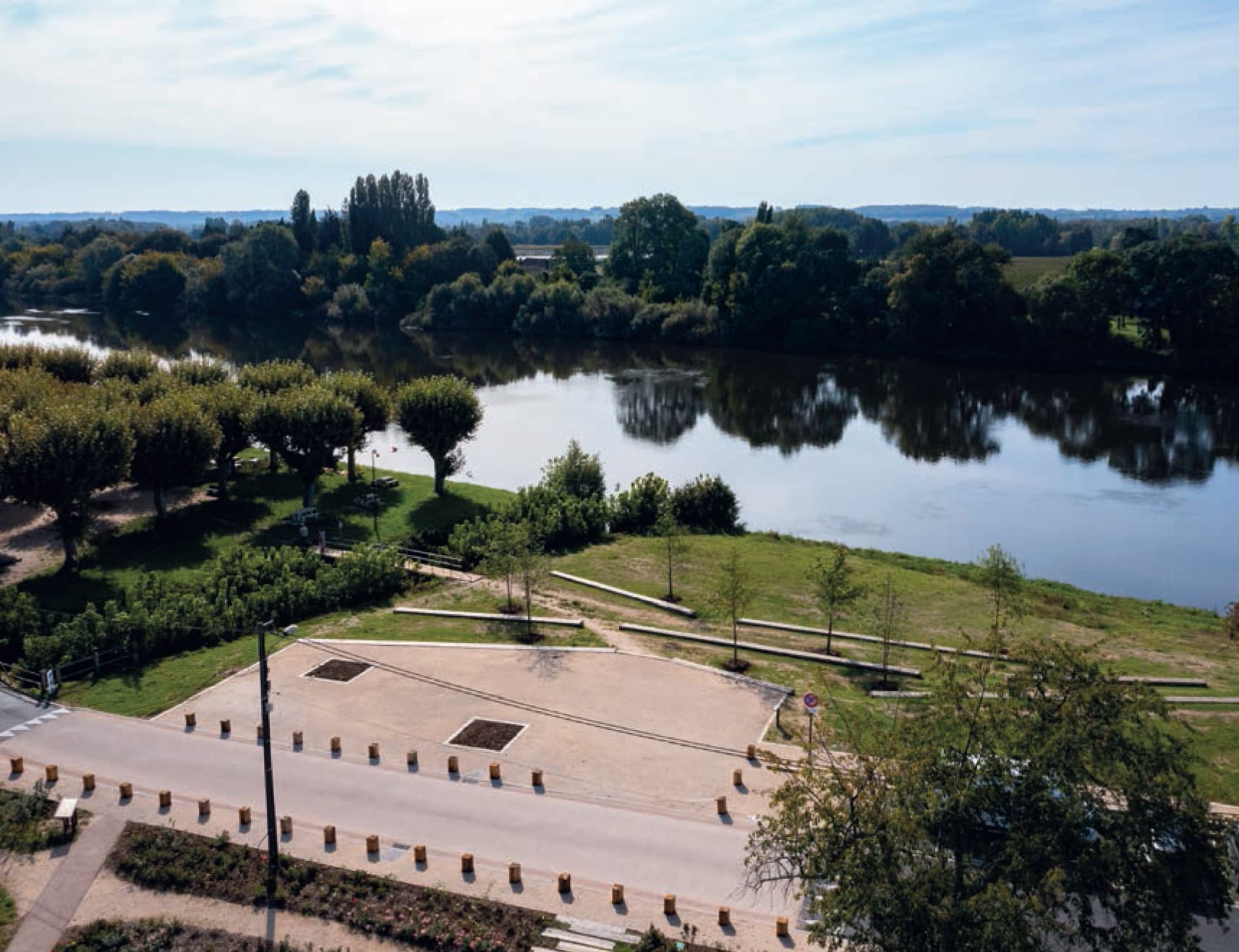 Aménagement paysager en bord de fleuve avec arbres, sentiers et reflets d'eau calme.