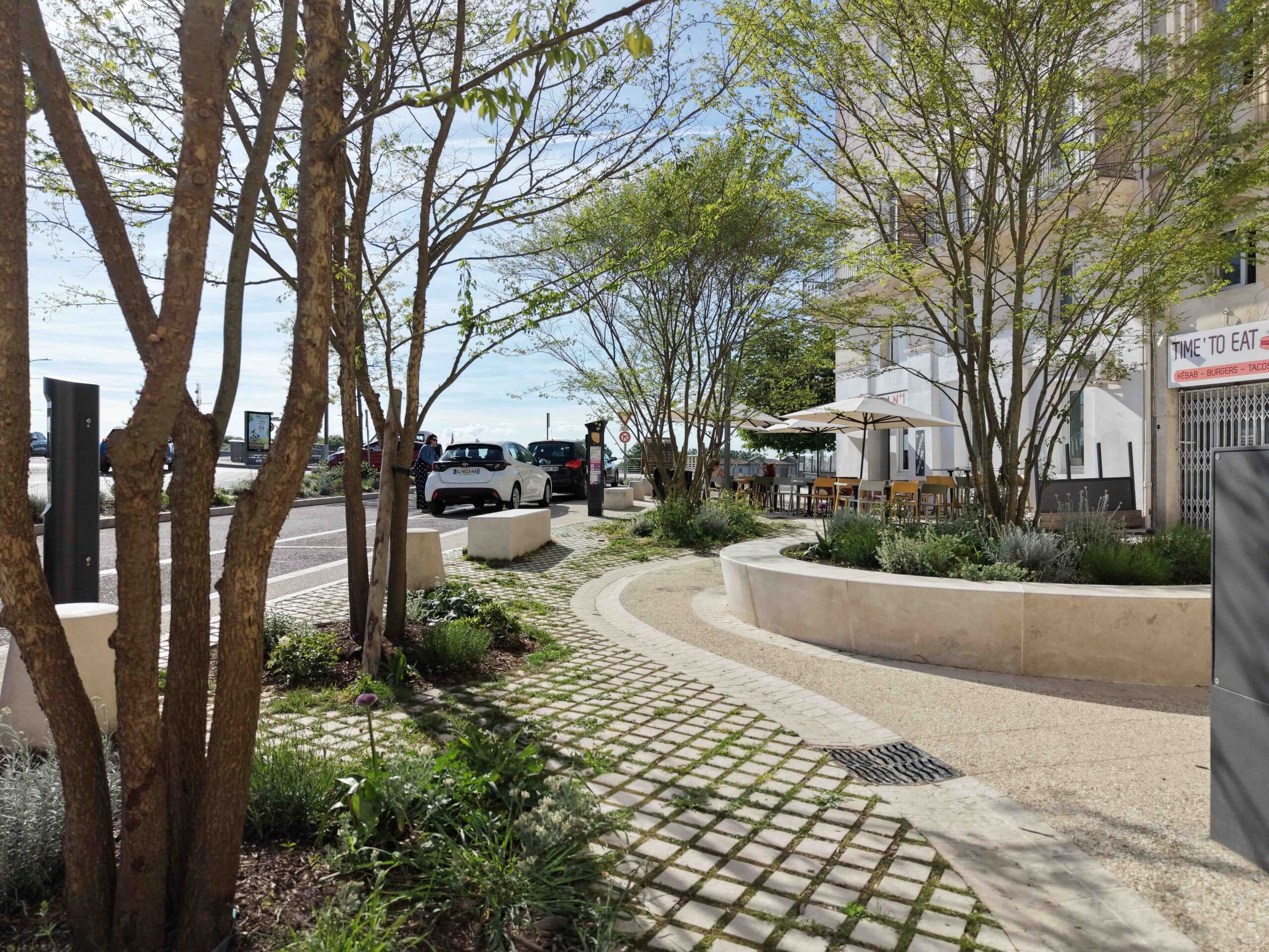 Terrasse TIME 'TO EAT, aménagement urbain végétalisé avec bancs en pierre et voitures garées sous le soleil.