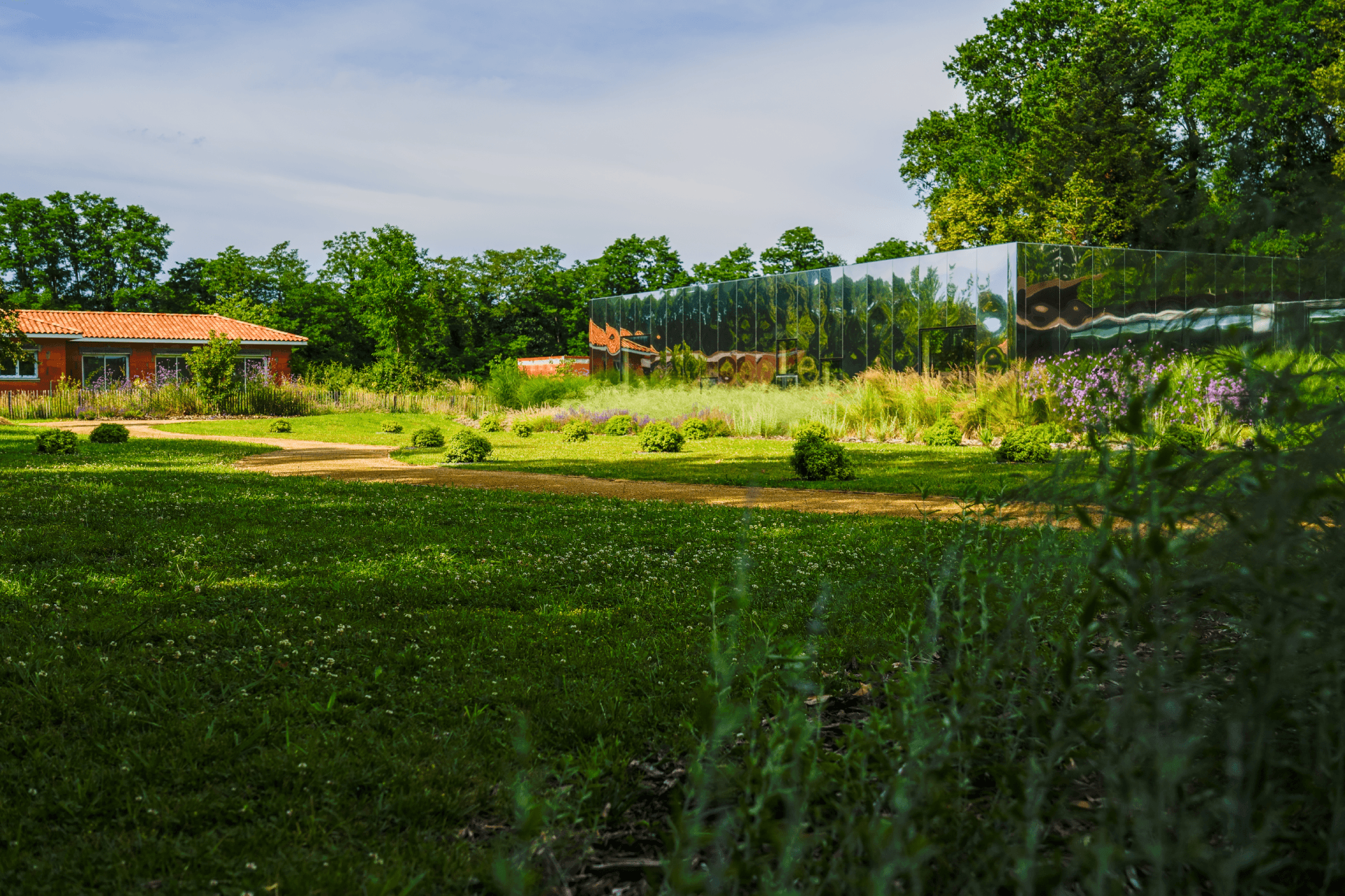 Jardin verdoyant avec bâtiment moderne en miroir reflétant les arbres et maison à toit de tuiles rouges en arrière-plan.