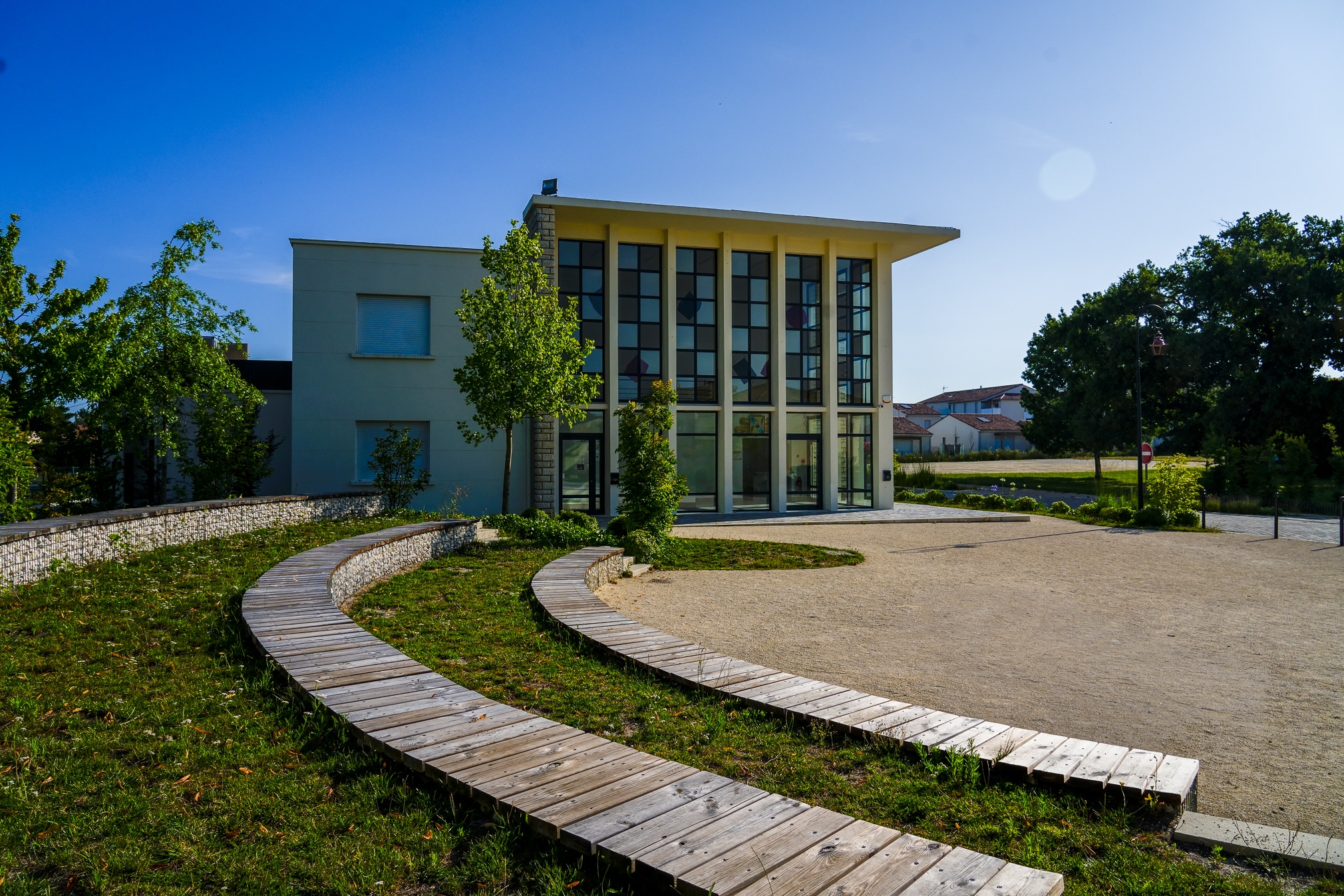 Bâtiment moderne à façade vitrée colorée avec sentiers en bois courbés et aménagement paysager sous un ciel bleu.