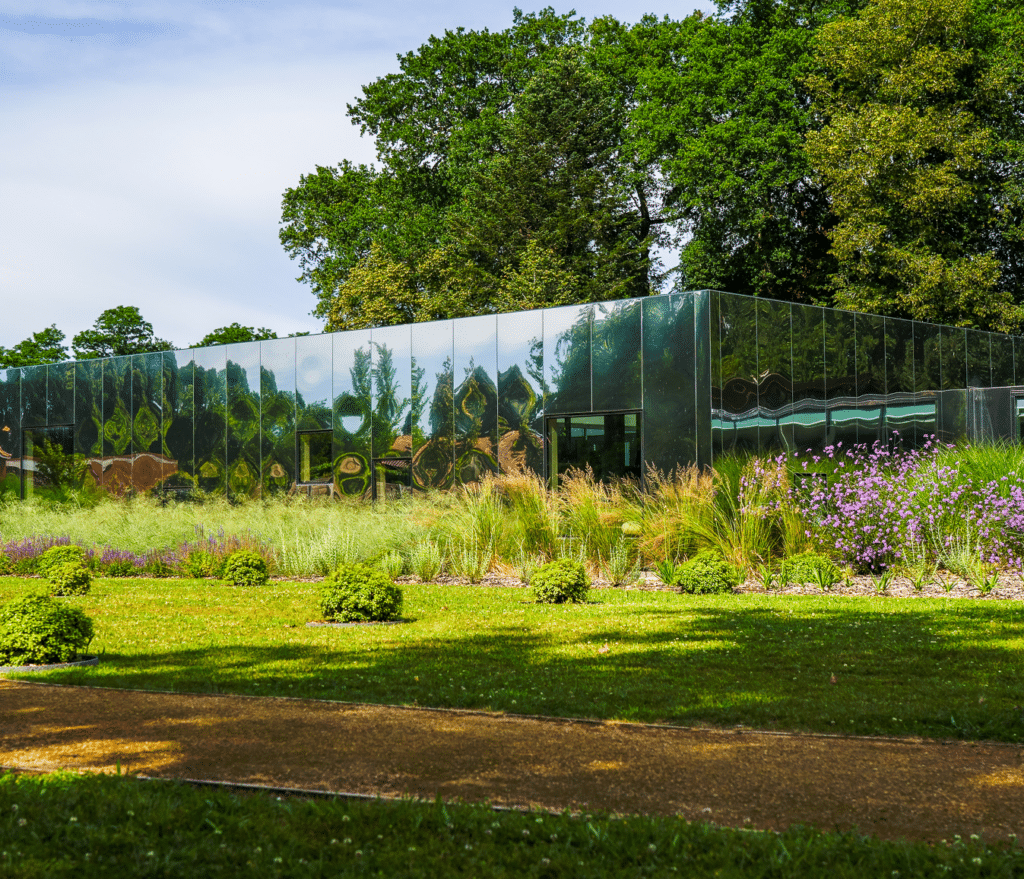 Architecture moderne : bâtiment miroir et jardin paysager Bâtiment à façade miroir reflétant des arbres, entouré d'un jardin fleuri et d'une pelouse ensoleillée.