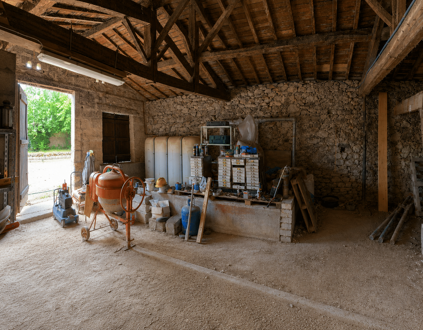 Intérieur d'une grange en pierre avec bétonnière orange, outils de construction et charpente en bois apparente.