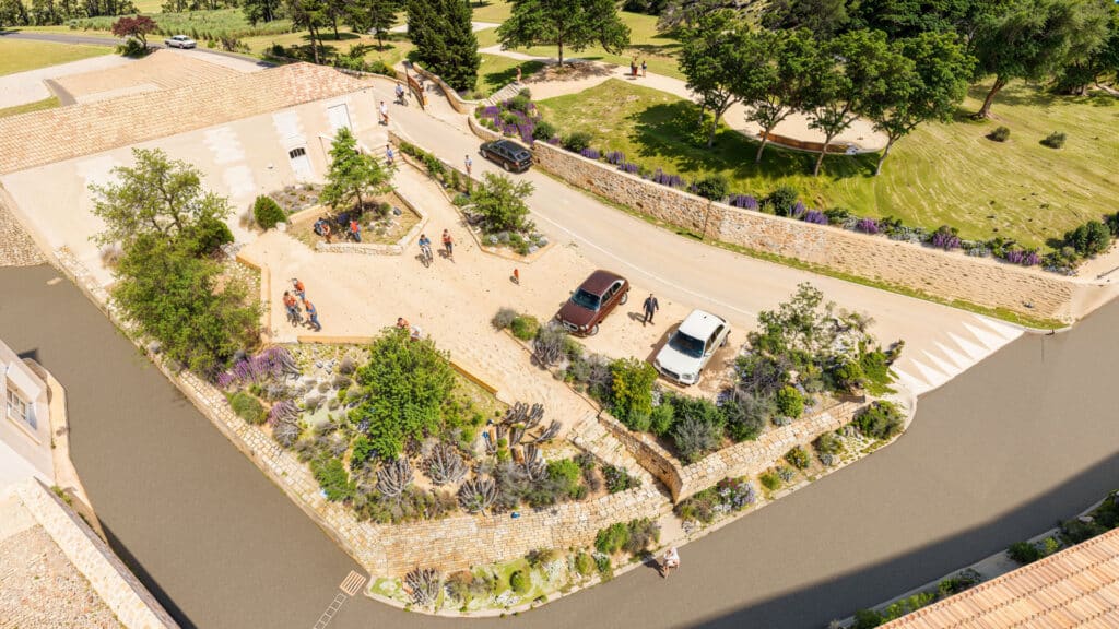 Domaine provençal et jardins paysagers vus du ciel Vue aérienne d'un domaine avec bâtiment en pierre, jardins fleuris, cyclistes et voitures stationnées.
