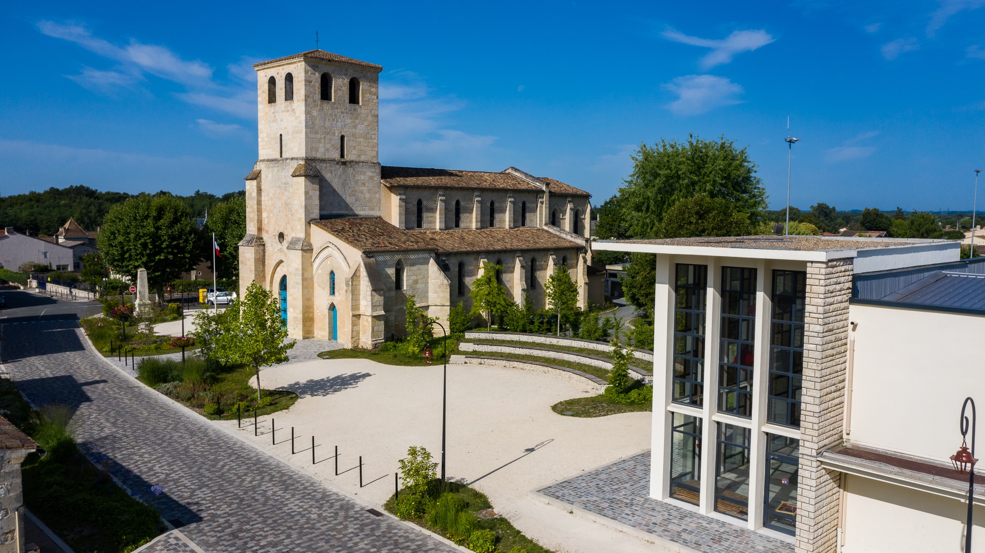 Place de village avec église en pierre et bâtiment moderne sous un ciel bleu.