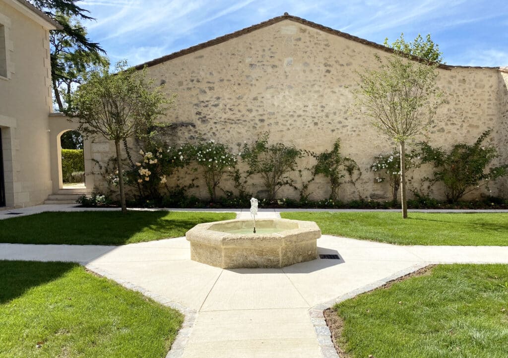 Fontaine de jardin octogonale et cour en pierre rustique Fontaine en pierre octogonale dans un jardin avec pelouse, sentiers blancs et grand mur de pierre rustique.