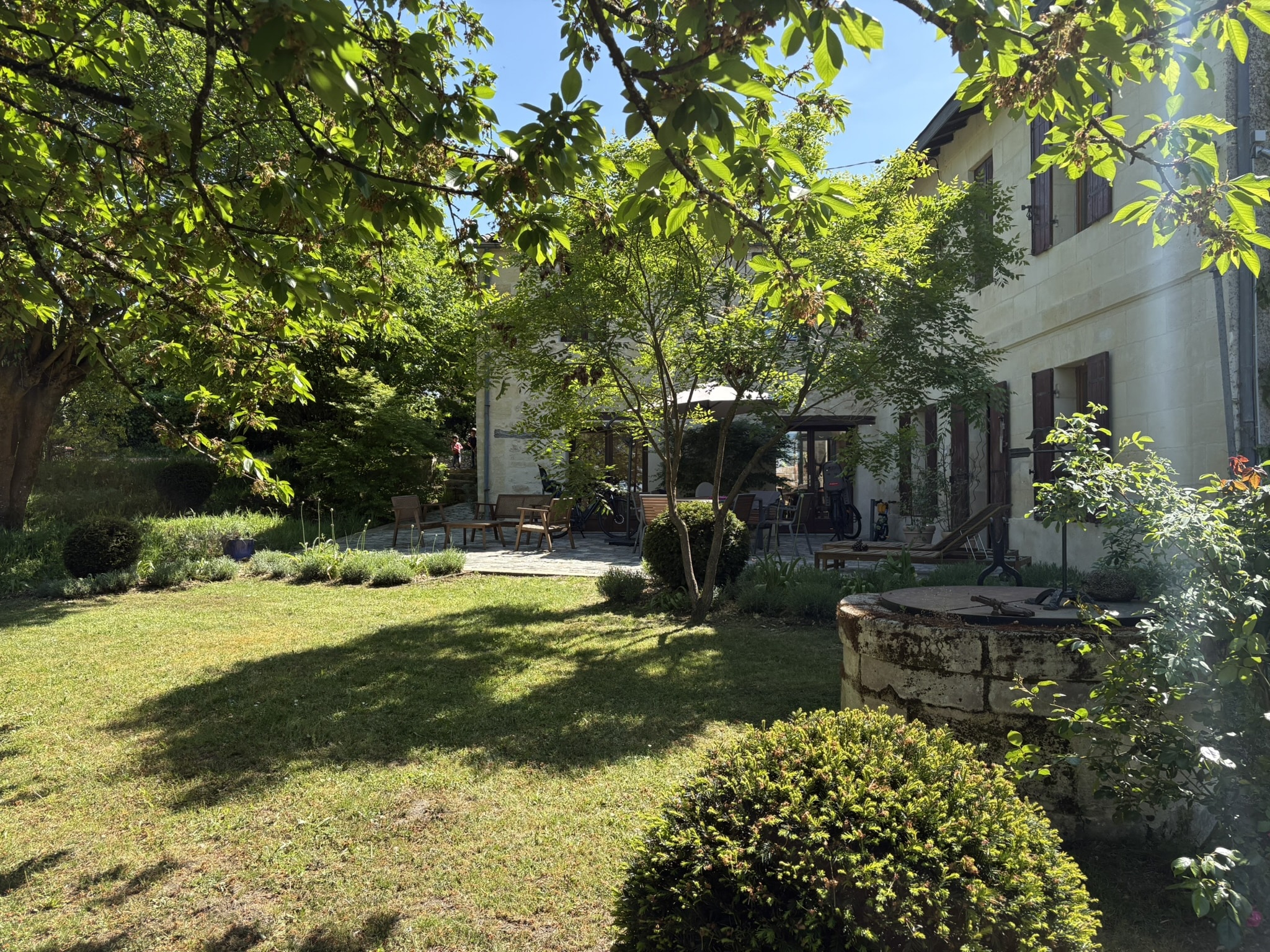 Jardin ensoleillé, terrasse de maison en pierre et puits ancien entourés de verdure luxuriante.
