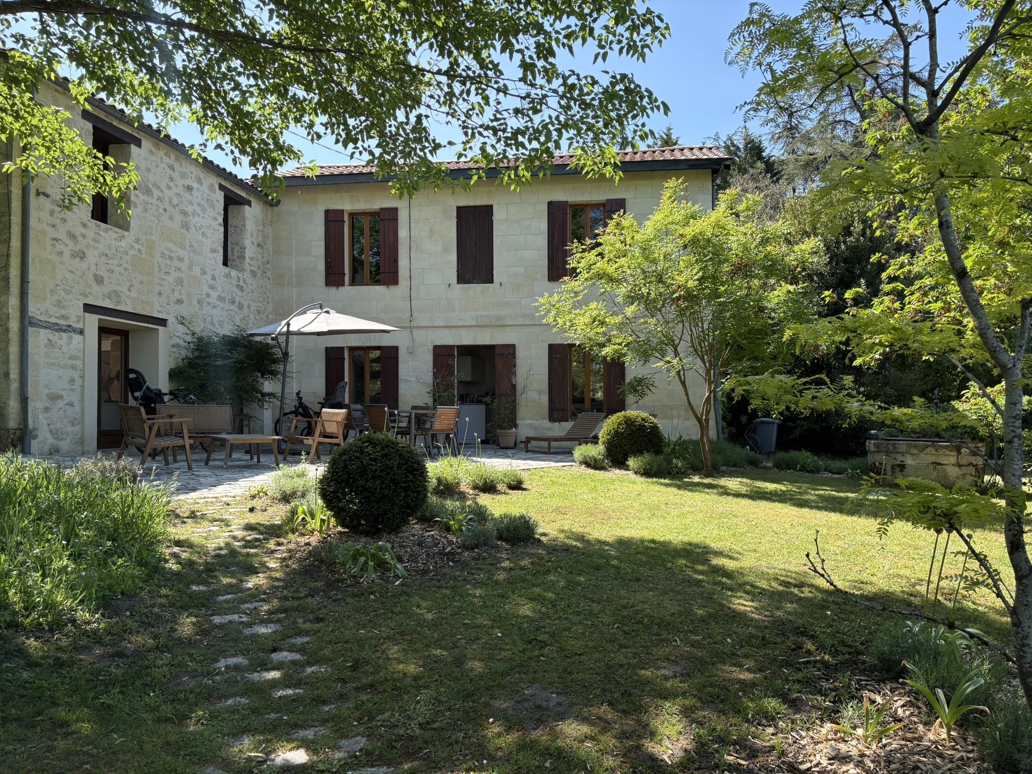 Maison en pierre avec volets bruns, terrasse meublée et jardin verdoyant sous un ciel bleu ensoleillé.