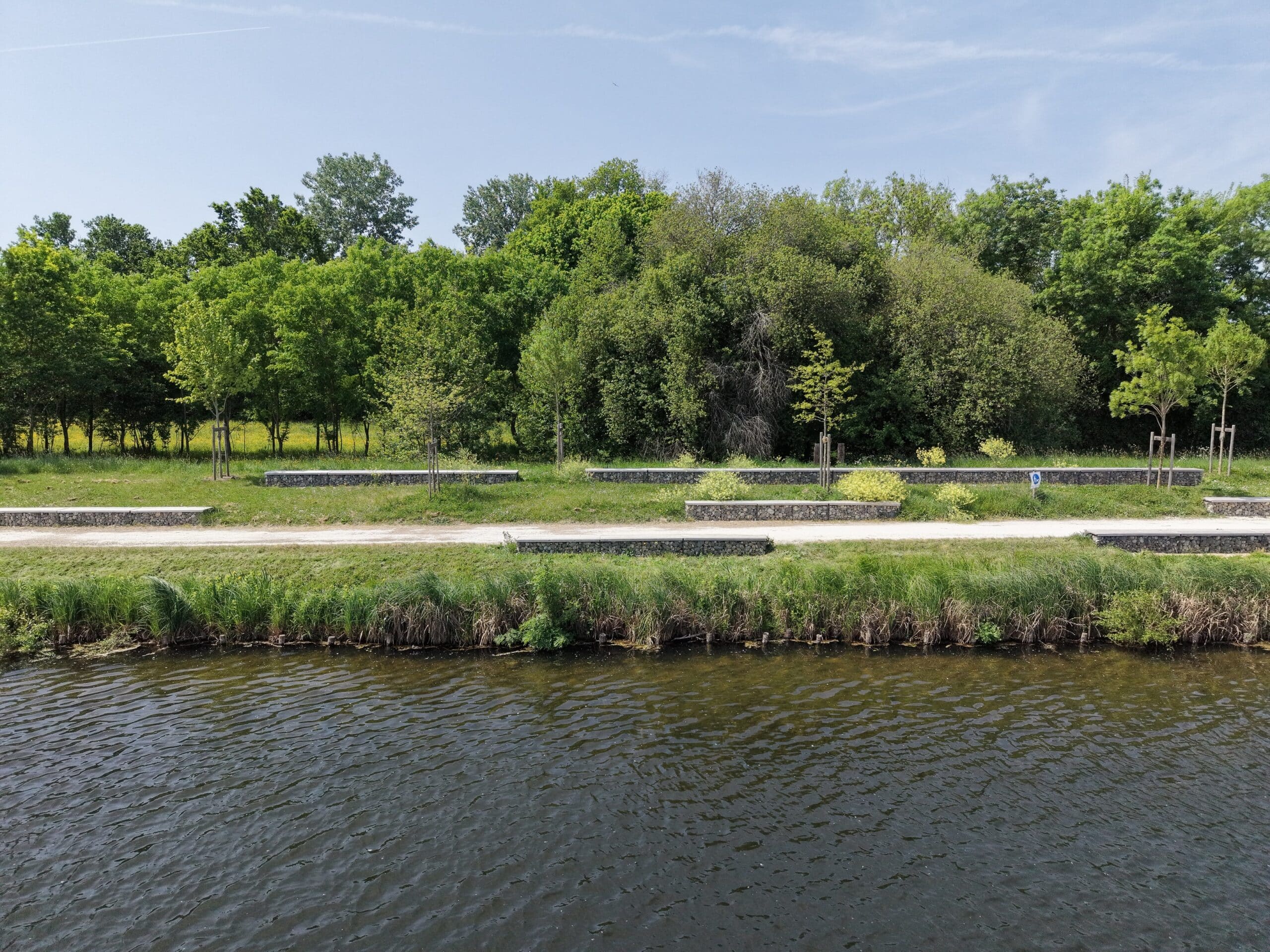 Bancs en gabion au bord d'un canal bordé par une forêt verdoyante sous un ciel bleu.
