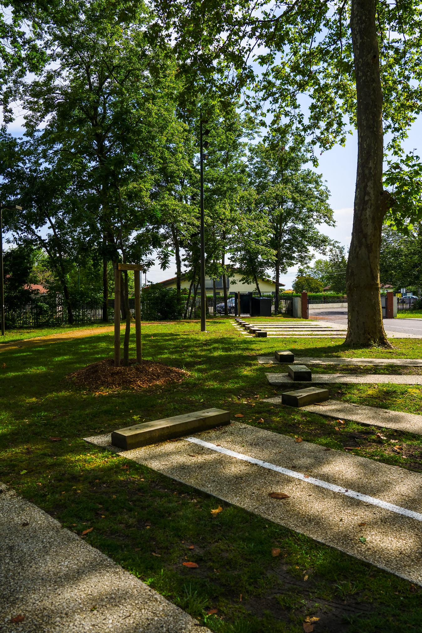 Parking paysager avec dalles de gravier, butoirs bois et arbres matures par temps ensoleillé.