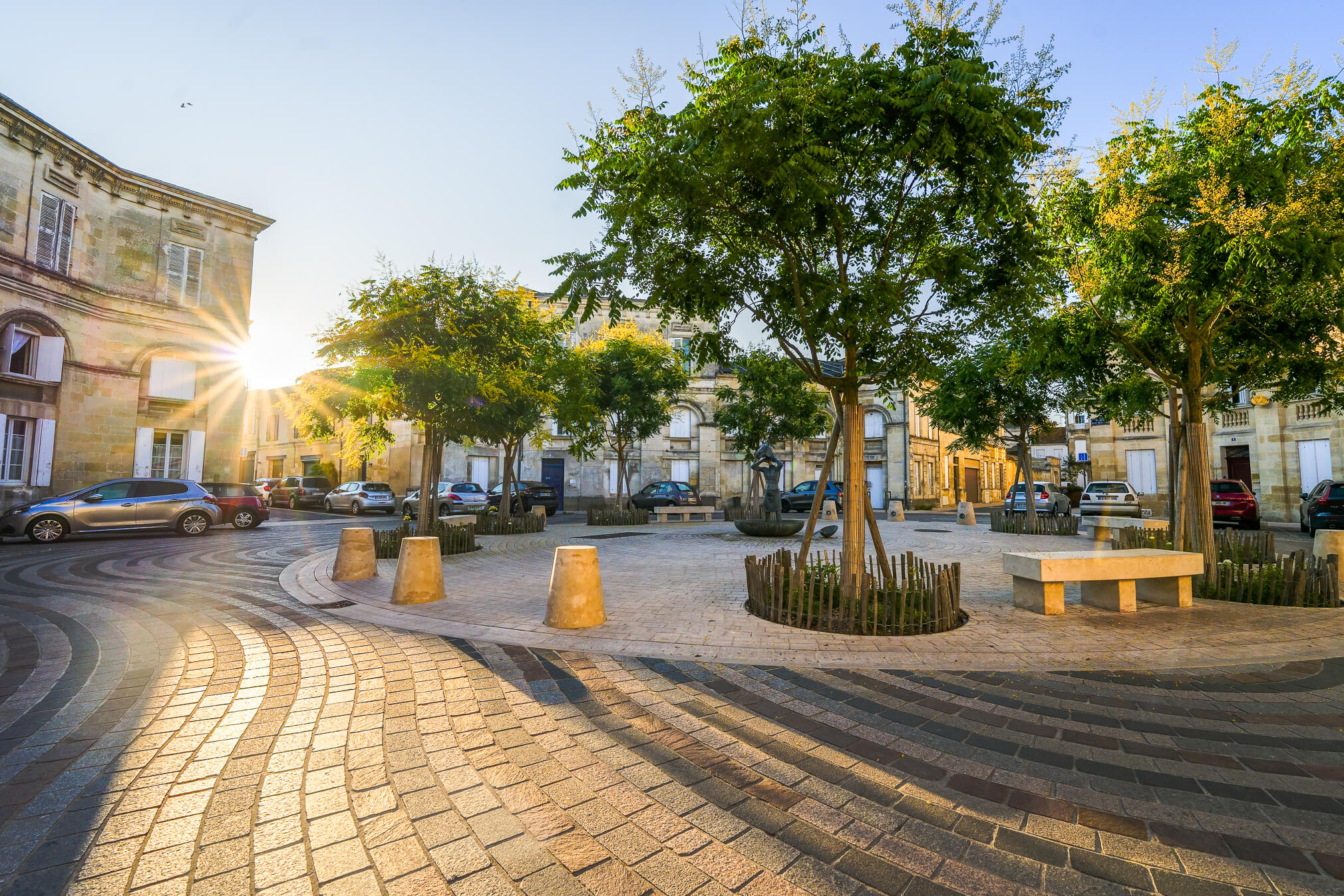 Place urbaine ensoleillée aux pavés ondulés, arbres et bâtiments anciens.