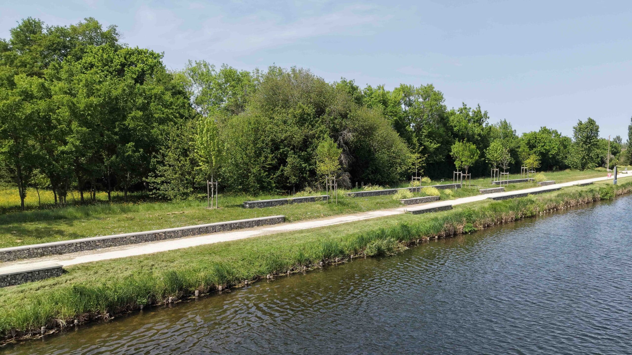 Sentier longeant un canal avec bancs en gabion et arbres sous un ciel clair.