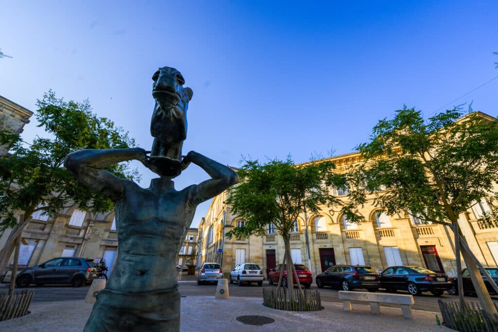 Statue d'homme à tête de cheval, Place Picard Bordeaux Statue en bronze d'un homme tenant une tête de cheval, Place Picard à Bordeaux, entourée de bâtiments classiques.