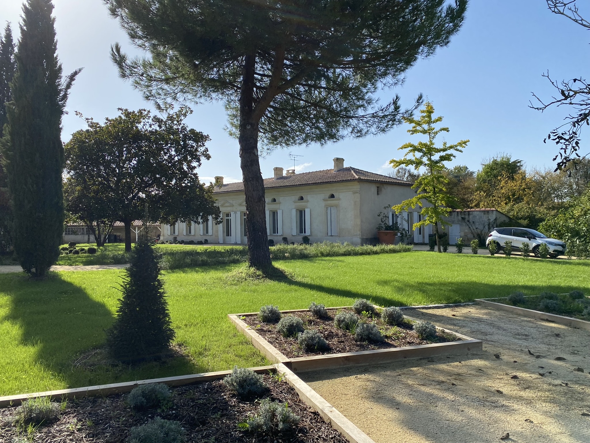 Villa méditerranéenne en pierre, volets blancs, grand jardin avec pins, parterres et pelouse sous ciel bleu.
