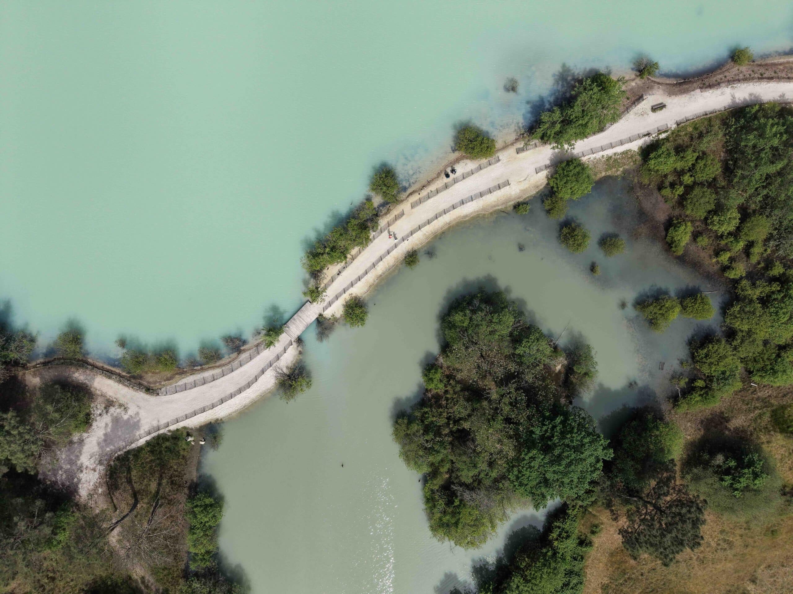 Sentier sinueux entre un lac turquoise et un étang verdoyant bordé de végétation, vue aérienne.