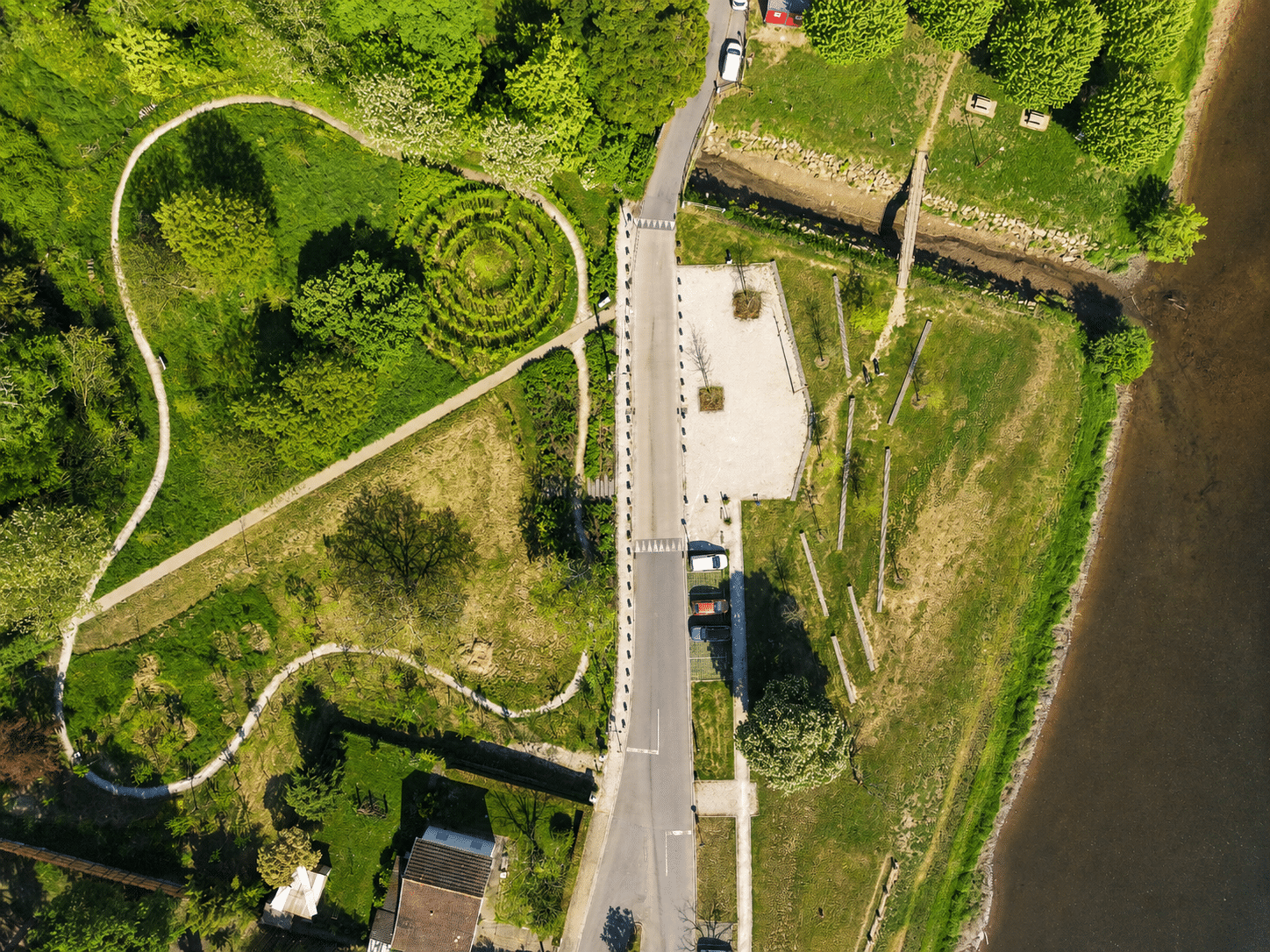 Parc avec sentier sinueux, labyrinthe végétal, route et rive boisée vus de dessus.