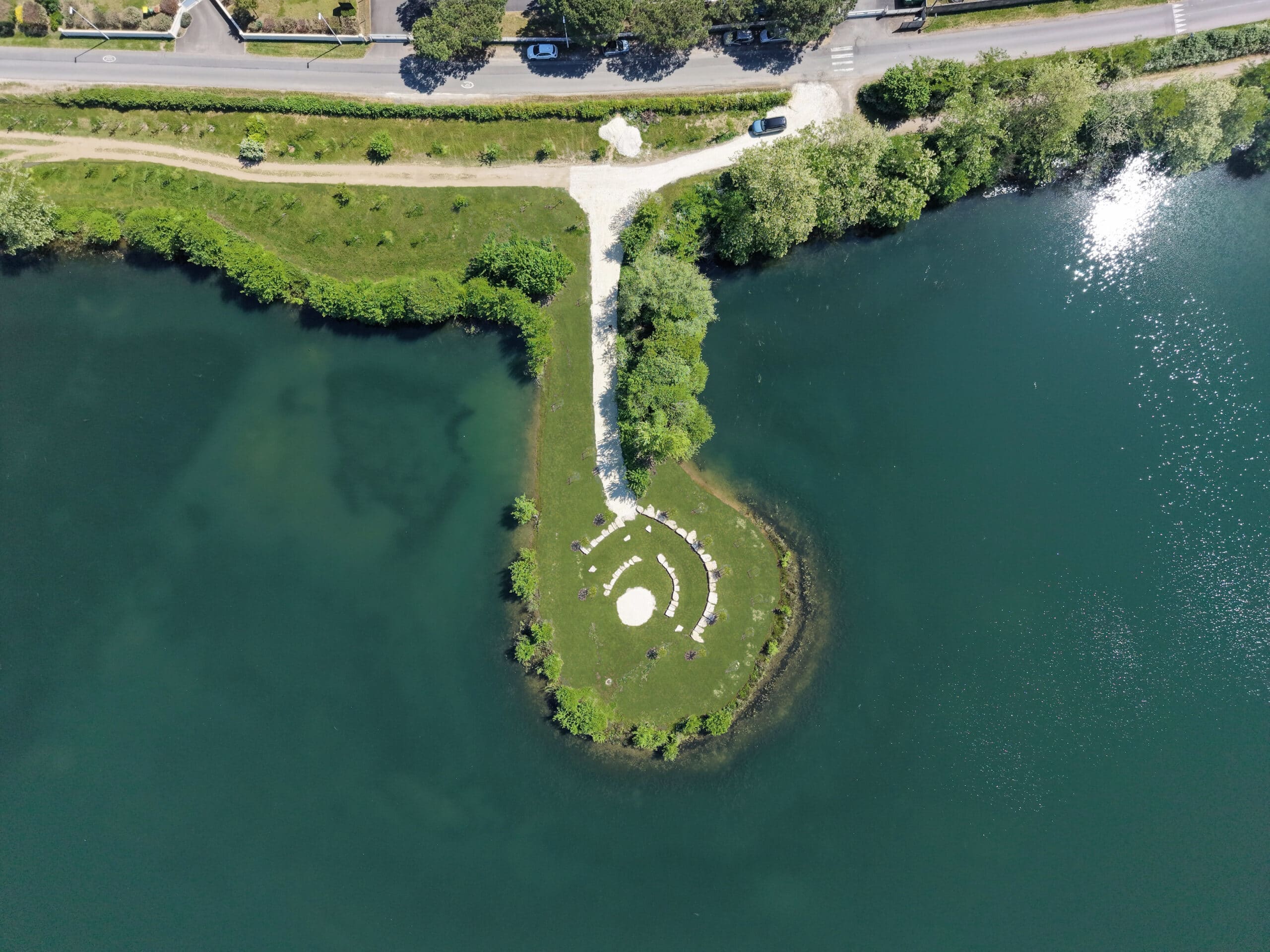 Péninsule circulaire avec cercle de pierres sur un lac turquoise, vue aérienne boisée.